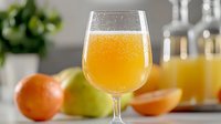 Fresh citrus juice in wine glass with orange and apple fruits in background, selective focus on sparkling beverage against soft blurred kitchen setting.