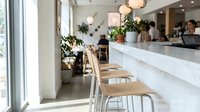 Modern minimalist cafe interior with white marble counter, wooden seats, pendant lighting, and potted plants near large windows, suitable for hospitality and design projects.