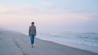 Young woman walking alone on sandy beach during sunset with gentle waves and pastel sky creating peaceful coastal atmosphere for wellness and travel concepts.