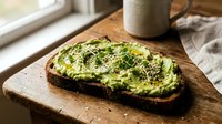 Avocado toast on dark bread with seeds and herbs on wooden cutting board, healthy breakfast meal with coffee mug in natural kitchen lighting.
