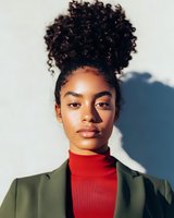 Young African American woman with natural curly hair in high bun wearing red turtleneck and olive green blazer against white wall with dramatic shadow.