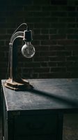 Vintage industrial desk lamp with exposed bulb on wooden table against dark brick wall background, suitable for interior design and decor projects.