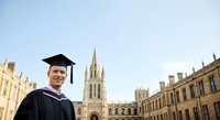 Graduate student wearing academic regalia and mortarboard cap smiling in front of historic university buildings and Gothic architecture, celebrating educational achievement and graduation ceremony.
