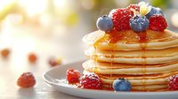 Stack of golden pancakes topped with fresh raspberries and blueberries on white plate, soft morning light creates warm breakfast atmosphere.