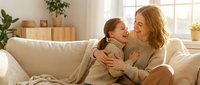 Happy mother and daughter embracing on couch in cozy living room with natural light streaming through window creating warm family bonding moment at home.