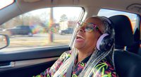 Joyful African American woman with dreadlocks wearing headphones and sunglasses laughing while sitting in car passenger seat during sunny day road trip.