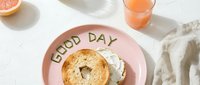 Toasted bagel with cream cheese on a pink plate with Good Day lettering, grapefruit juice, and linen napkin on a white surface.