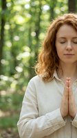 Peaceful middle-aged Caucasian woman with curly red hair meditating outdoors in natural forest setting with hands in prayer position and eyes closed.