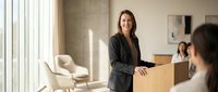 Professional businesswoman in dark suit holding cardboard box in modern office during corporate relocation or job transition with natural lighting.