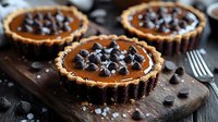 Chocolate caramel tarts with crispy shortbread crust topped with dark chocolate chips on rustic wooden board, homemade dessert photography.