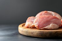 Fresh raw chicken breast fillets on round wooden cutting board against dark background, selective focus with copy space for culinary designs.