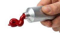 Hand squeezing a metal tube of red paint, with a swirling curl of vivid crimson paint emerging from the tip on white background.