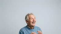 Senior man with gray hair and light skin tone, wearing blue sweater, smiling and gesturing while speaking against neutral background for portrait and communication concepts.