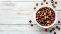 Chocolate chips and colorful candy pieces in white ceramic bowl on rustic wooden background with scattered morsels, top view with copy space.