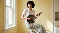 Young woman with curly hair playing banjo while sitting on windowsill in cozy home interior with natural lighting for music and lifestyle content.