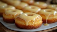Fresh baked vanilla glazed donuts with creamy caramel topping dripping down sides on white ceramic plate, close up view showing soft texture and golden color.