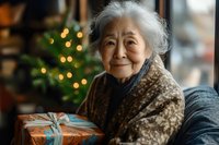 Senior Asian woman smiling warmly while holding wrapped Christmas present, wearing cozy leopard print cardigan against blurred festive lights and decorated tree background.