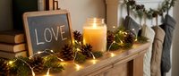 Cozy Christmas fireplace mantel with glowing candle, pine cones, fairy lights, fir branches, chalkboard with love inscription, and stockings.