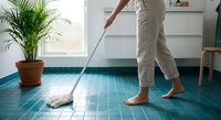 Woman mopping blue tile floor in modern kitchen with white cabinets and palm plant. Household cleaning and home maintenance concept.
