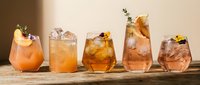 Five artisanal cocktails with amber and orange hues garnished with herbs and fruits arranged on wooden surface against warm beige background for bar menu.