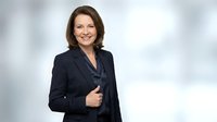 Confident middle-aged woman with shoulder-length brown hair, Caucasian ethnicity, wearing dark navy blazer over burgundy top, smiling at camera with hand on jacket. Corporate professional portrait.