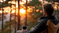 Young man with backpack enjoying warm coffee while watching golden sunset through forest trees during outdoor hiking adventure.