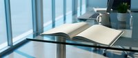 Open book on glass table by window with natural light streaming in, modern office or study space setup for reading and learning.