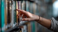 Female hand reaching for book on library shelf, bracelet on wrist, selective focus on fingers against blurred bookshelf background creates atmospheric depth.
