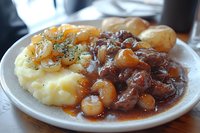 Tender beef stew with caramelized pearl onions served over creamy mashed potatoes and garnished with fresh herbs on white plate.