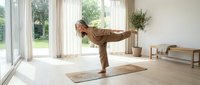 Mature man practicing yoga in bright modern living room with wooden floors and garden view, stretching in warrior pose on mat.
