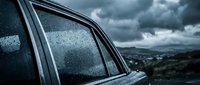 Car window with raindrops against dramatic stormy sky and landscape background creating moody atmospheric scene for weather and travel concepts.
