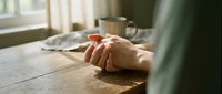 Hands clasped together on a rustic wooden table near a window, with a metal mug in the soft background light.