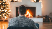 Man in gray sweater sitting near fireplace with decorated Christmas tree and warm lights creating cozy holiday atmosphere at home. Peaceful winter evening scene.