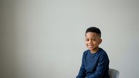 Young African American boy wearing navy blue long-sleeve shirt, seated against neutral gray background, smiling at camera with confident expression, suitable for educational, family, and lifestyle content.