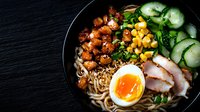 Asian noodle bowl with glazed chicken, soft-boiled egg, cucumber, corn, and ramen noodles served on black plate, dramatic lighting creates appetizing mood.