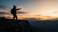 Young male hiker standing on rocky mountain peak at sunset, pointing toward distant valley landscape. Silhouetted figure wearing backpack against golden sky with clouds and mountain ranges below.