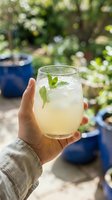Hand holding glass of refreshing lemonade with fresh mint leaves and ice cubes outdoors in garden setting, perfect for beverage, summer drink, and lifestyle content.