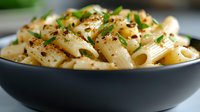 Creamy penne pasta in black ceramic bowl garnished with fresh herbs, red pepper flakes and black pepper. Close-up view shows texture of sauce coating pasta.