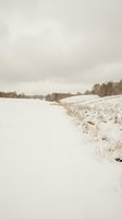 Snowy winter landscape with frozen lake and bare trees under overcast sky creating peaceful rural scenery for seasonal backgrounds and nature designs.