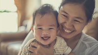 Asian mother and laughing baby share joyful moment at home. Natural window light creates warm atmosphere for authentic family lifestyle portrait.