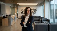 Professional businesswoman with crossed arms standing confidently in modern corporate office lobby with contemporary furniture and large windows.