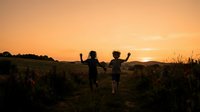 Two children running through a wildflower field at golden hour sunset, arms raised in joy and freedom, celebrating nature and outdoor adventure during summer evening.