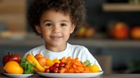 Happy mixed-race toddler boy with curly hair smiling at camera while sitting at kitchen table with colorful plate of fresh fruits and vegetables for healthy eating.