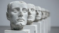 Ancient marble death masks arranged in linear perspective, showing detailed facial features on white pedestals against gray background. Historical sculpture collection.