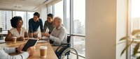 Diverse team of professionals collaborating in modern office conference room with city skyline view, discussing business strategy using laptop and tablet during daytime meeting.