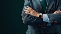 Confident business professional in dark suit with crossed arms and wristwatch against green background, portraying leadership and authority in corporate setting.