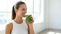Young woman with brown hair, Caucasian ethnicity, wearing white sleeveless top, drinking fresh green juice or smoothie in bright modern interior space with natural window light.