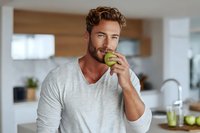 Young Caucasian man with beard eating green apple in modern kitchen, promoting healthy lifestyle and nutrition habits.