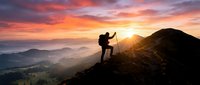 Hiker silhouette celebrating success on mountain peak during golden sunrise with dramatic orange sky and layered mountain ranges in misty valley below.