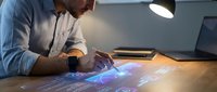 Young Caucasian businessman in blue shirt working with holographic data visualization interface at modern office desk with laptop and smartphone.